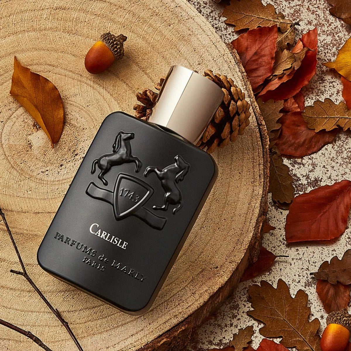 Parfums de Marly Carlisle Eau de Parfum bottle on a natural wood slice surrounded by autumn leaves, pinecones and acorns.