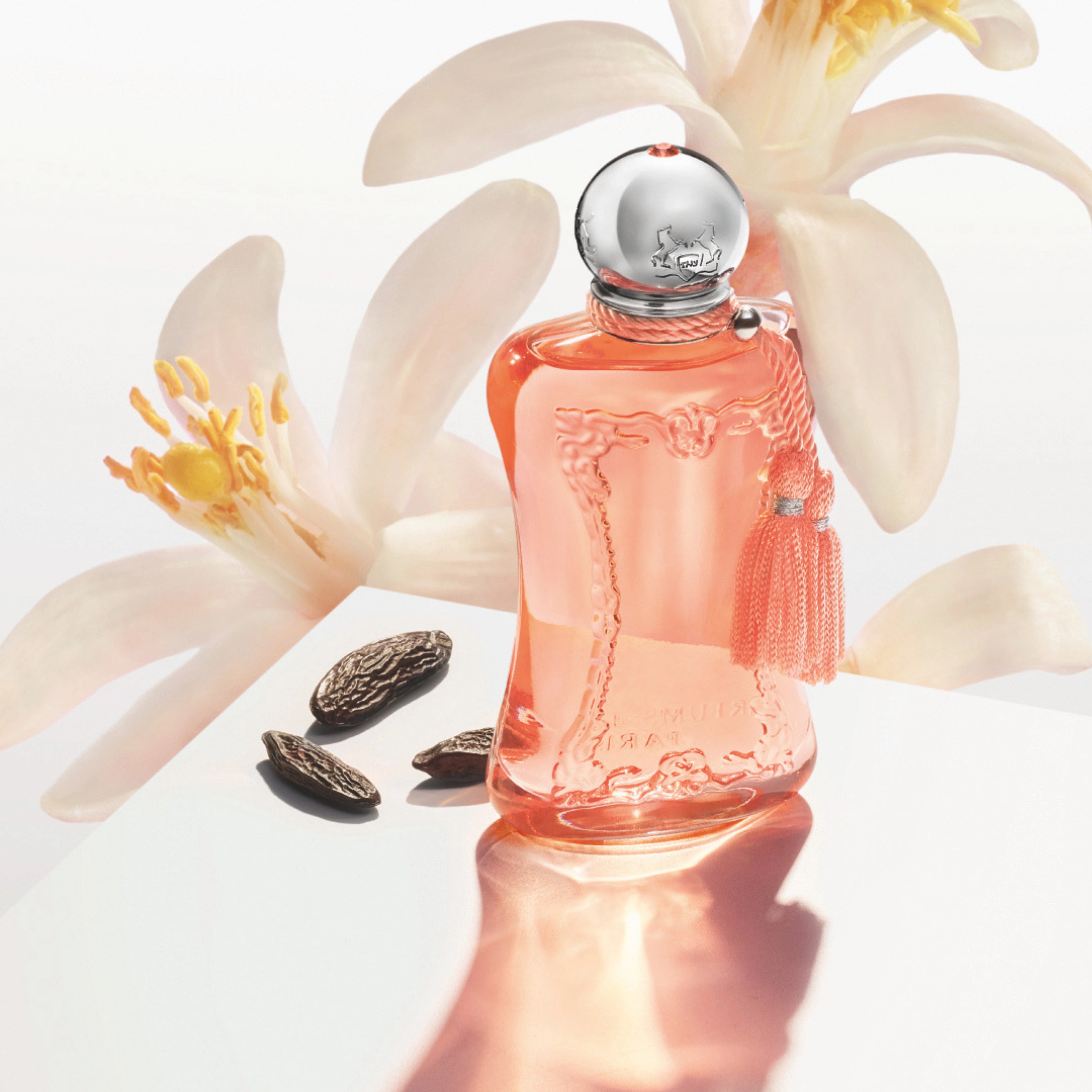 Parfums de Marly Athénaïs Eau de Parfum bottle with a translucent coral finish, silver cap and matching coral tassels displayed beside tonka beans with white blossoms in the background.