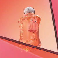 Parfums de Marly Athénaïs Eau de Parfum bottle with a translucent coral finish, silver cap and matching coral tassels displayed against a coral gradient background with reflective panels.