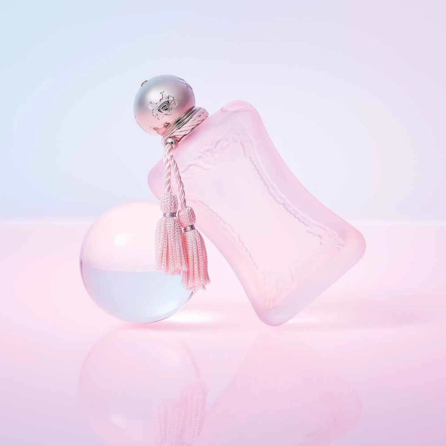 Parfums de Marly Delina La Rosée Eau de Parfum bottle with a frosted soft pink finish, silver cap and matching pale pink tassels displayed on a white sphere against a pastel pink background.