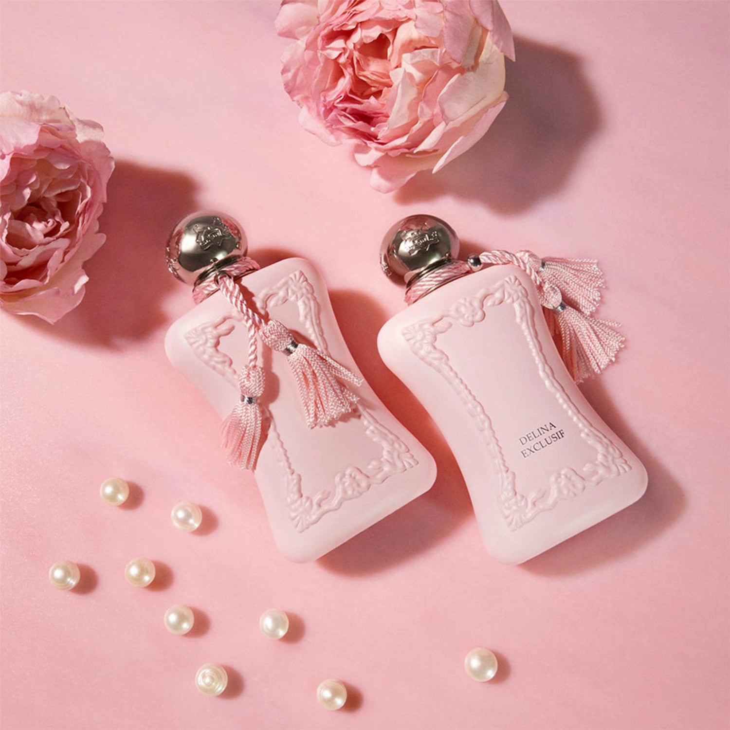 Parfums de Marly Delina Exclusif Eau de Parfum bottles with silver caps and pink tassels displayed on a pink background with blooming pink roses and scattered pearls.