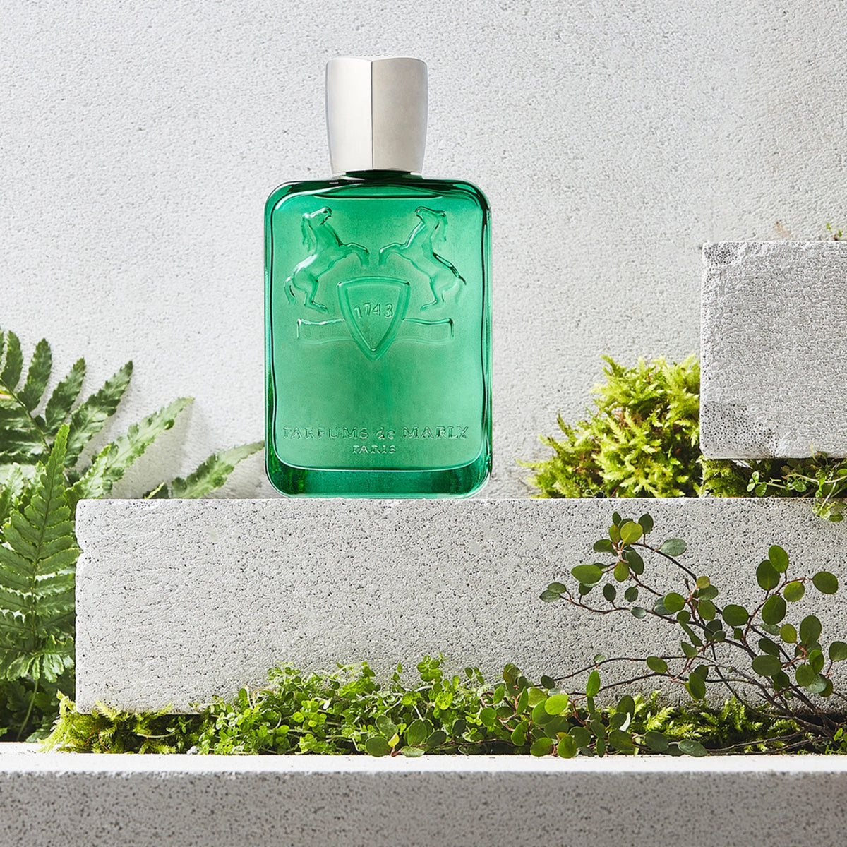 Parfums de Marly Greenley Eau de Parfum bottle in translucent emerald-green glass with an embossed horse crest and silver cap displayed among lush green foliage on textured stone plinths in natural daylight.