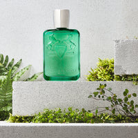 Parfums de Marly Greenley Eau de Parfum bottle in translucent emerald-green glass with an embossed horse crest and silver cap displayed among lush green foliage on textured stone plinths in natural daylight.