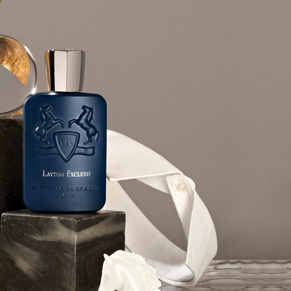 Parfums de Marly Layton Exclusif Eau de Parfum bottle in deep matte midnight-blue with an embossed horse crest and silver cap displayed on black marble blocks beside a crisp white dress shirt, creating a refined luxury setting.
