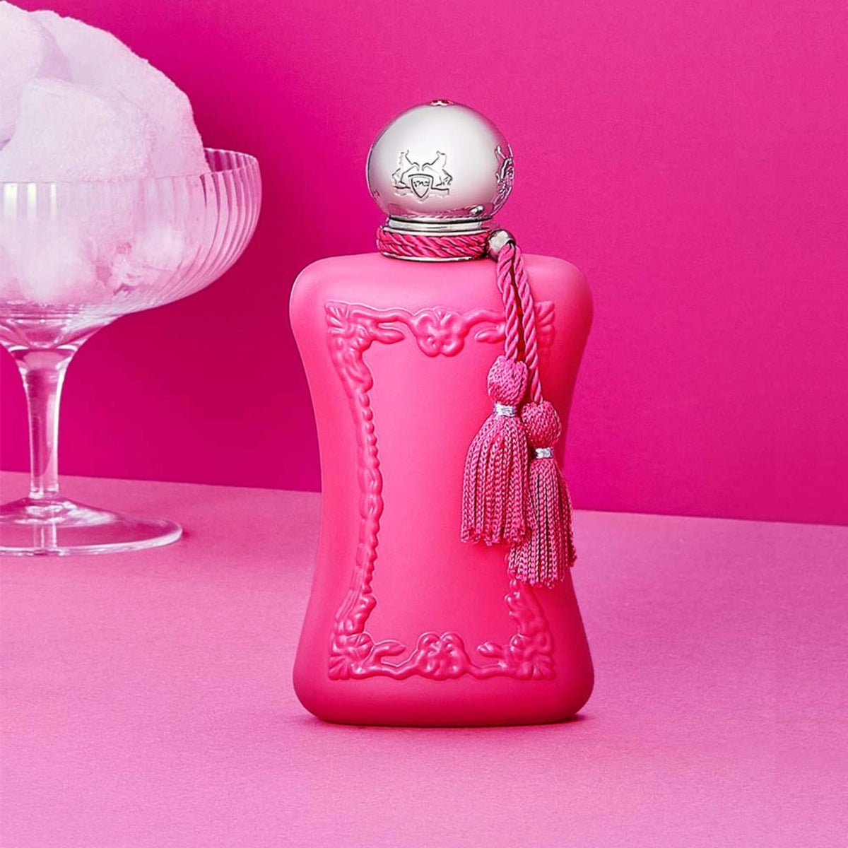 Parfums de Marly Oriana Eau de Parfum bottle in matte raspberry pink with an ornate embossed frame, silver cap and matching tassels displayed beside a coupe glass filled with marshmallows against a vibrant pink backdrop.
