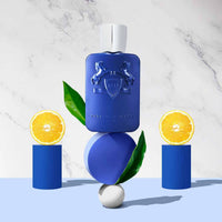 Parfums de Marly Percival Eau de Parfum bottle with a matte royal blue finish and silver cap displayed with orange slices and green leaves on blue geometric podiums.