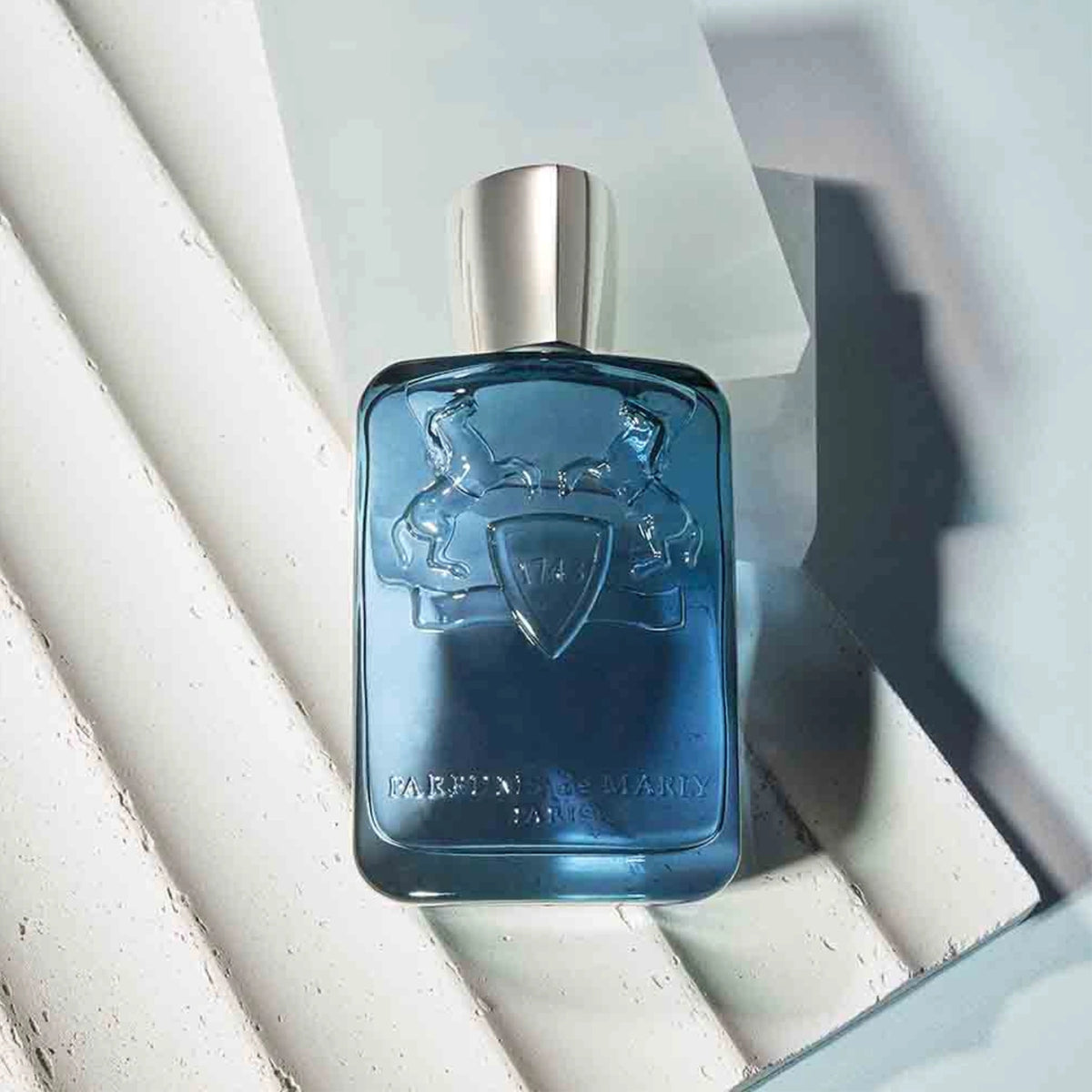 Parfums de Marly Sedley Eau de Parfum bottle with a translucent steel blue finish, silver cap and embossed horse crest displayed on white architectural surfaces.